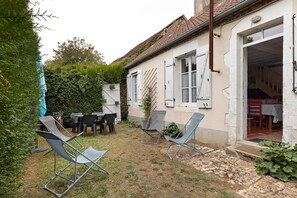 A rustic outdoor seating area with metal chairs and a table, surrounded by a garden and a building with a tiled roof.