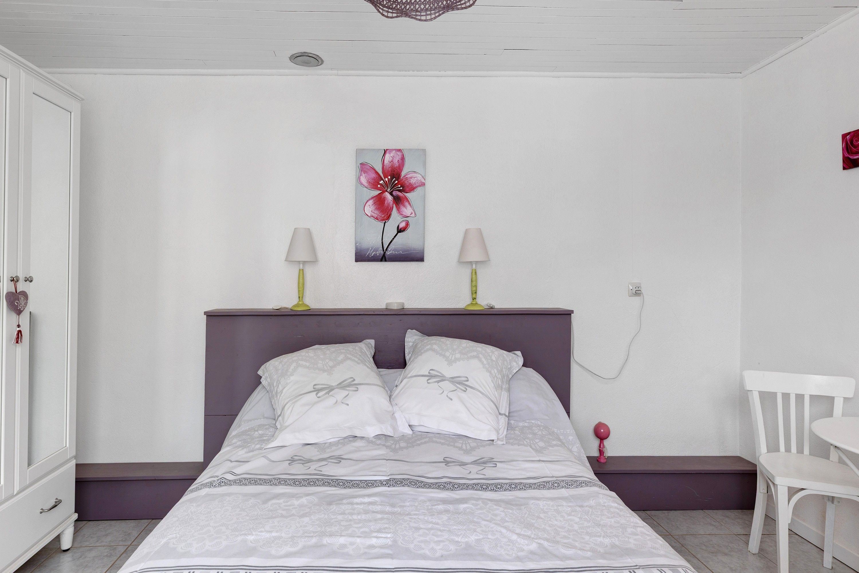 Une chambre à coucher avec un lit, deux lampes de chevet, un tableau accroché au mur, une chaise blanche et une armoire blanche.