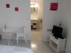 Une chambre d’hôtel avec un lit, une petite table ronde sur laquelle se trouve un vase, une chaise blanche, un téléviseur posé sur un meuble et une salle de bain visible à travers une porte ouverte.
