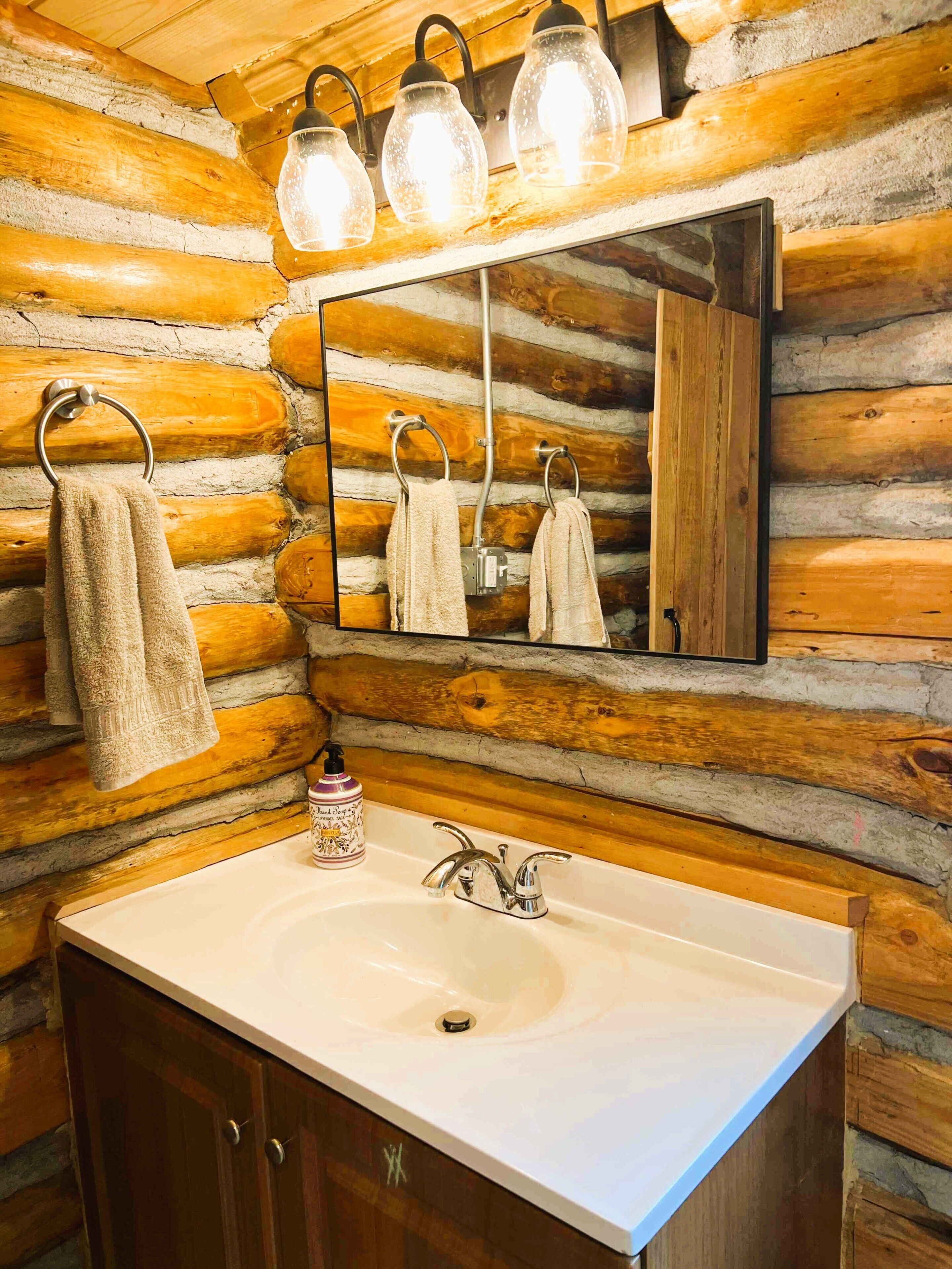A bathroom with a wooden log wall, a sink with a mirror, and three hanging light fixtures.