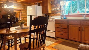 A wooden cabin interior with a dining table, chairs, a kitchen area with a refrigerator, stove, and cabinets, and a window with a view of trees. - Historic 1896 Cabin Retreat on 70-Acre Private Forest (Raymond)