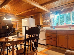 A wooden cabin interior with a dining table, chairs, a kitchen area with a refrigerator, stove, and cabinets, and a window with a view of trees.