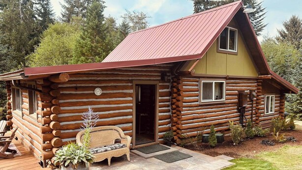 A log cabin with a red roof, a front porch with a bench, and a small garden area. - Historic 1896 Cabin Retreat on 70-Acre Private Forest (Raymond)