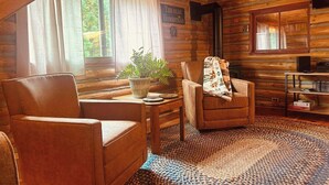 A cozy log cabin interior with a sofa, armchair, potted plant, and a fireplace. - Historic 1896 Cabin Retreat on 70-Acre Private Forest (Raymond)