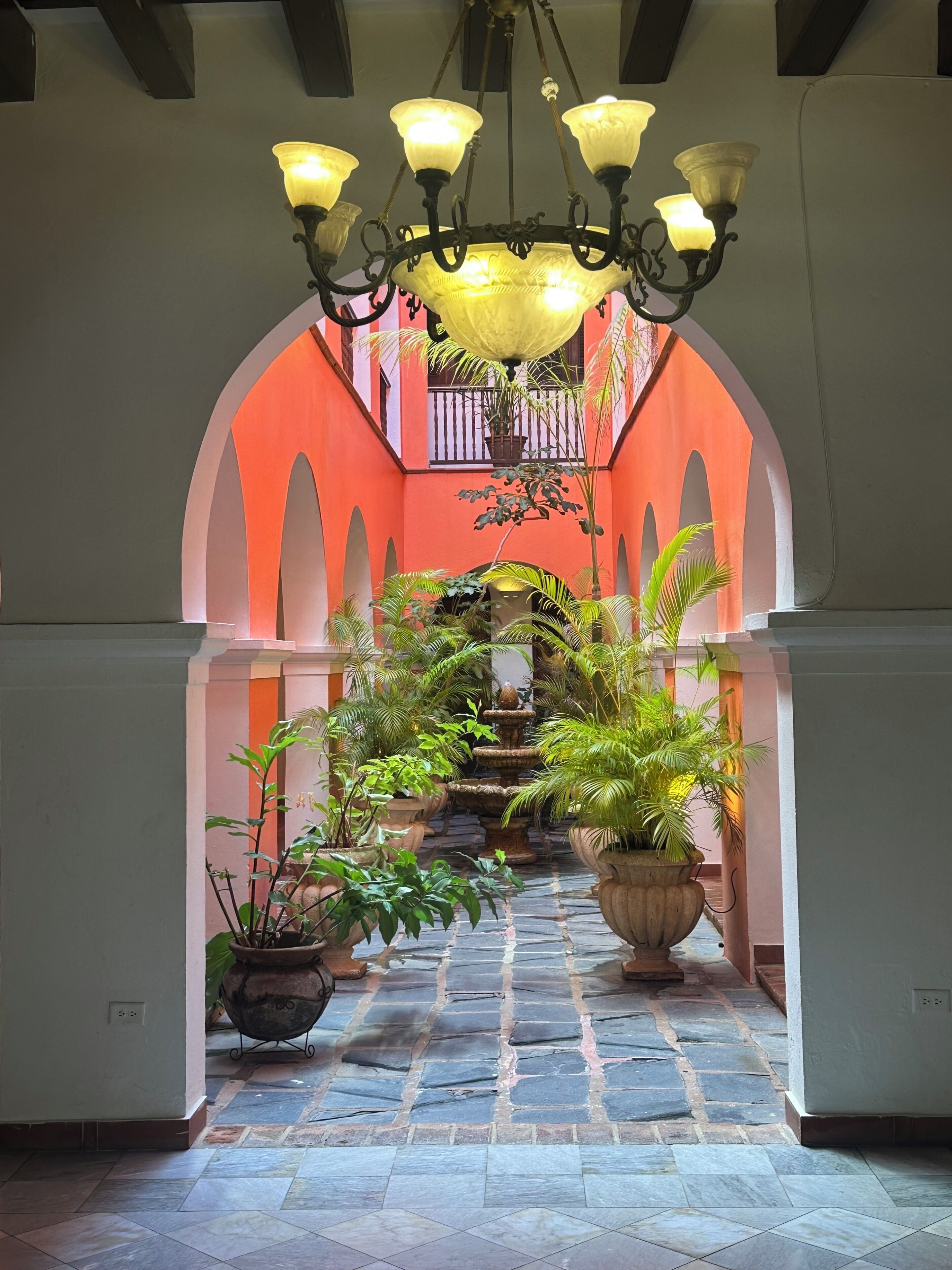 Un corredor de piedra con un estanque central, flanqueado por macetas con plantas y un candelabro ornamentado.