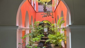 Un couloir dallé de pierre, avec une fontaine centrale, bordé de plantes en pot et surmonté d’un lustre somptueux.
