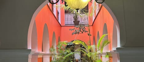 Un corredor de piedra con un estanque central, flanqueado por macetas con plantas y un candelabro ornamentado.