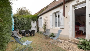 Un'area salotto esterna rustica con sedie in metallo e un tavolo, circondata da un giardino e un edificio con tetto in tegole.