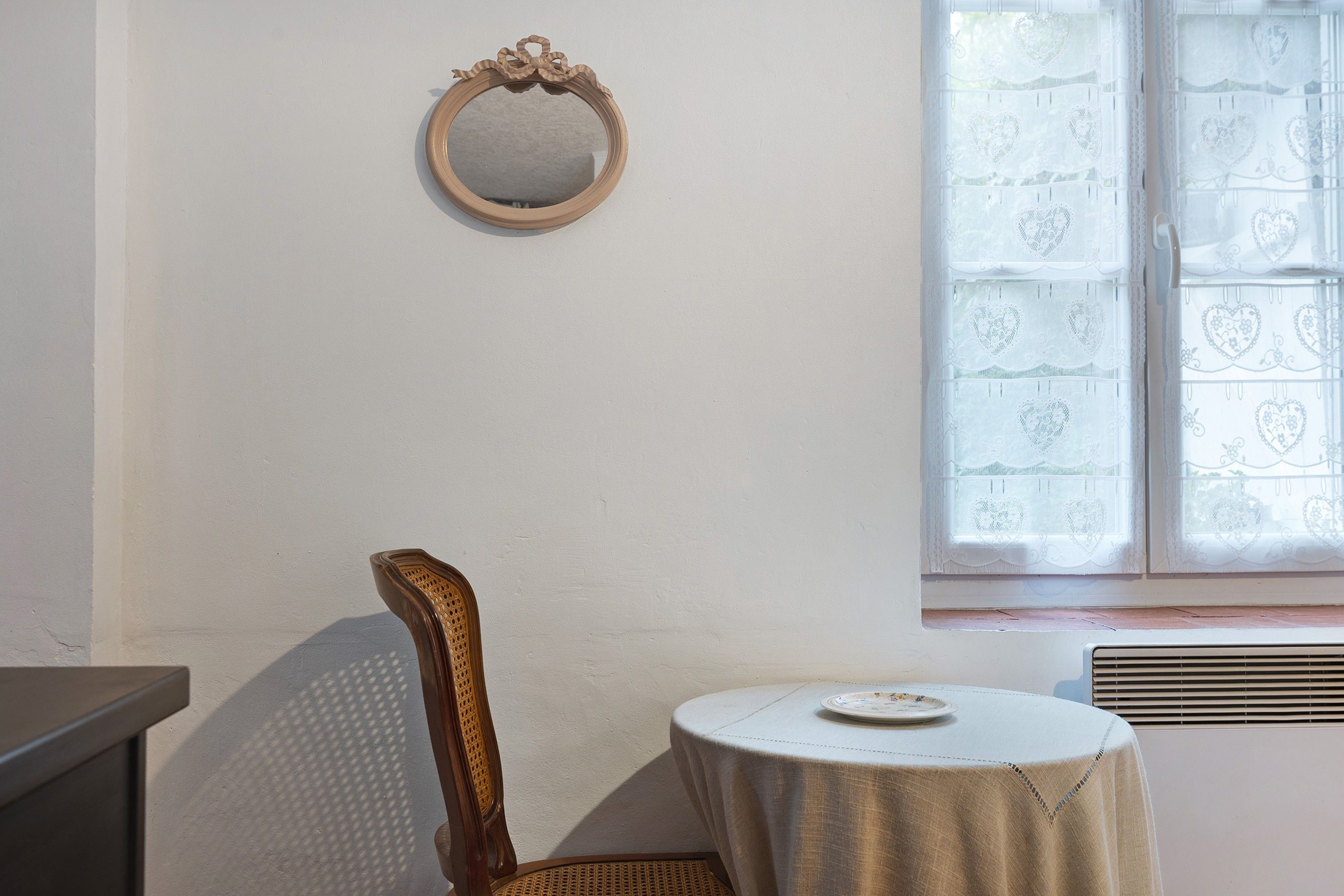 Een kamer met een ronde spiegel aan de muur, een stoel, een klein rond tafeltje met een bord en een raam met kantgordijnen.