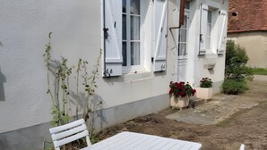 Ein weißes Haus mit Garten-Terrasse, inklusive weißem Tisch und Stühlen, einem Fenster mit Fensterläden und einem kleinen Blumenbeet mit roten Blüten.