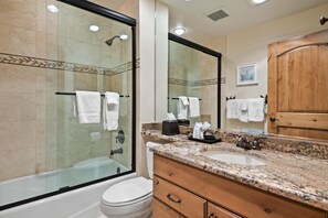 Ein modernes Badezimmer mit einer gläsernen Duschkabine, einer weißen Toilette, einem Waschbecken mit Granitplatte, Holzschränken und einem großen Spiegel.