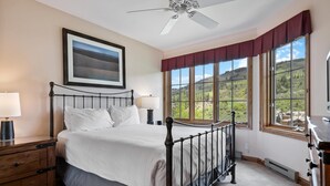 Un dormitorio con cama, ventilador de techo, ventana con vistas al exterior y mesita de noche con lámpara.