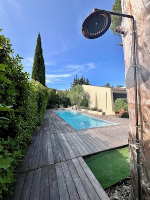 Une terrasse en bois mène à une piscine rectangulaire dotée d’une douche, le tout étant entouré de verdure et d’un grand arbre.