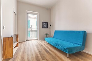 Een kamer met een houten vloer, een blauwe bank, een kleine tafel met een vaas en een deur naar een balkon.