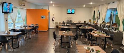 A modern dining area with multiple tables and chairs, a kitchen counter with food displays, and two televisions mounted on the walls.