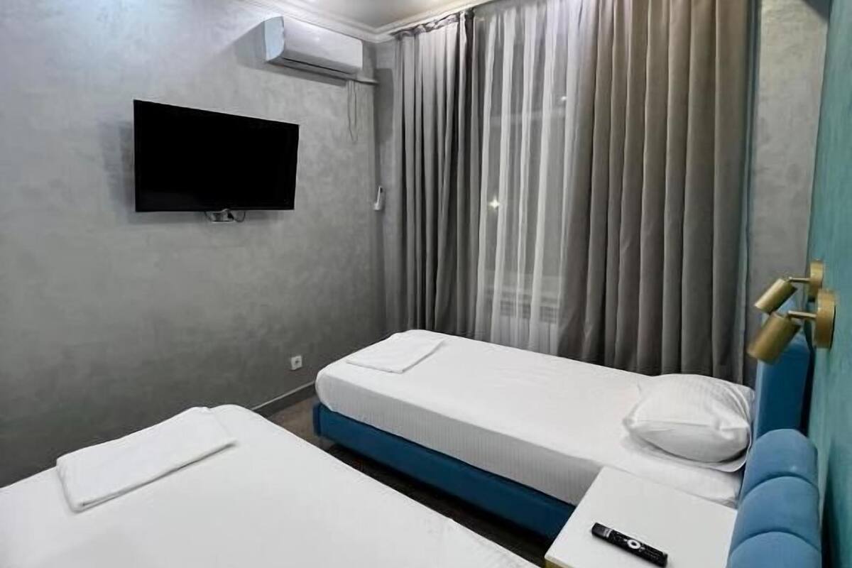 A hotel room with two single beds, a TV mounted on the wall, an air conditioning unit, and a curtain.