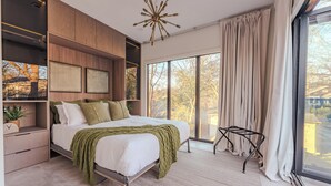 Ein modernes Schlafzimmer mit einem großen Bett, integrierten Stauraumlösungen, einer besonderen Deckenleuchte und Blick durch große Fenster.