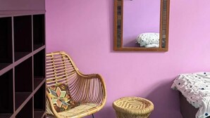 Huoneessa on violetti seinä, rottinkituoli, pieni punottu jakkara, peili ja valkoisella peitteellä varustettu sänky.