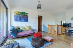 Een woonkamer met een bank, kleurrijke kussens, een schilderij, een lamp en een trap.