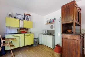 Een kleine keuken met gele kastjes, een houten stoel, een witte koelkast en een plank met diverse spullen.