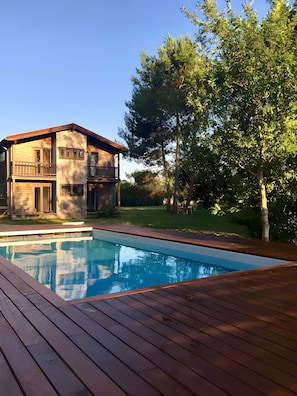Una casa moderna con piscina, terraza de madera y un gran árbol en el patio trasero.
