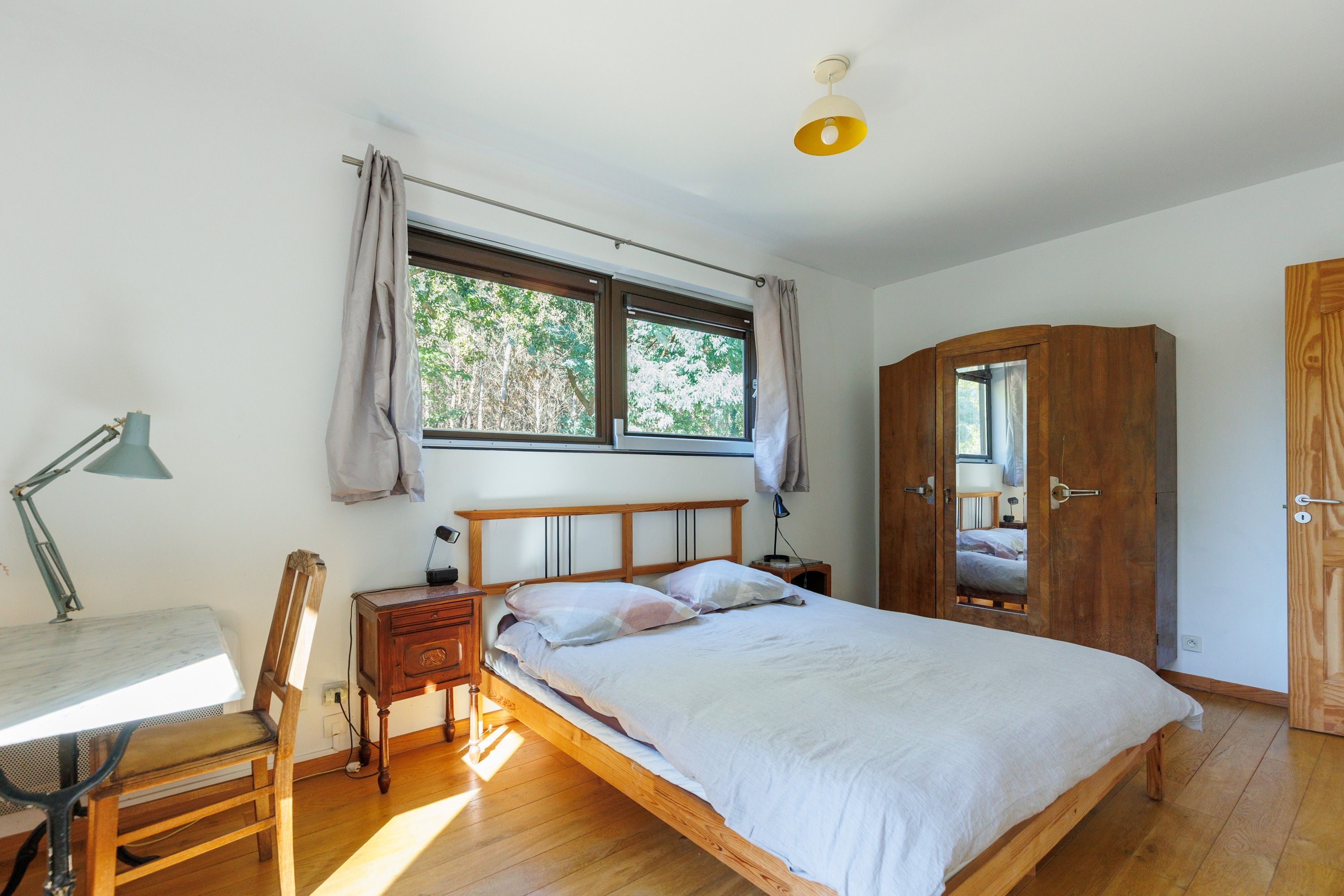 Een slaapkamer met een houten bed, een bureau met lamp, een kledingkast en een raam met gordijnen.