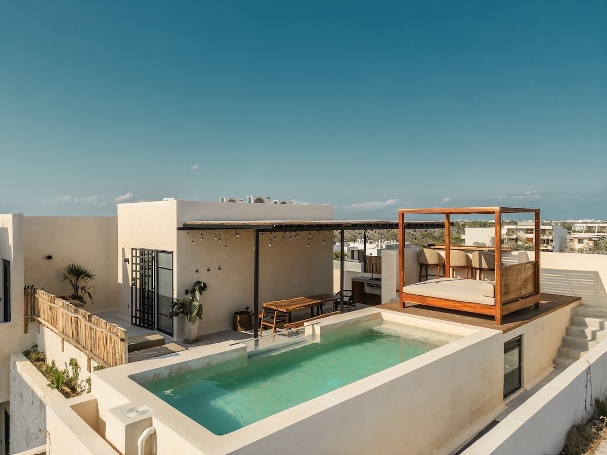 A rooftop pool with a wooden deck, a covered seating area, and a view of the cityscape.