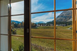 Blick durch ein Fenster mit Gittermuster, zeigt eine gebirgige Landschaft mit Gebäuden und blauem Himmel.