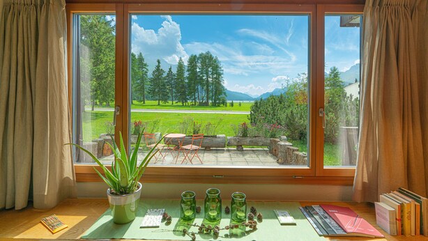 Ein Zimmer mit großem Fenster, Holzboden, einer Topfpflanze und einem Tisch mit Büchern und einer Vase.