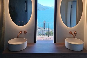 Ein Badezimmer mit zwei Waschbecken, jeweils mit einem Kupferarmaturen, und einem großen Fenster mit Blick auf ein Gewässer und eine Berglandschaft.