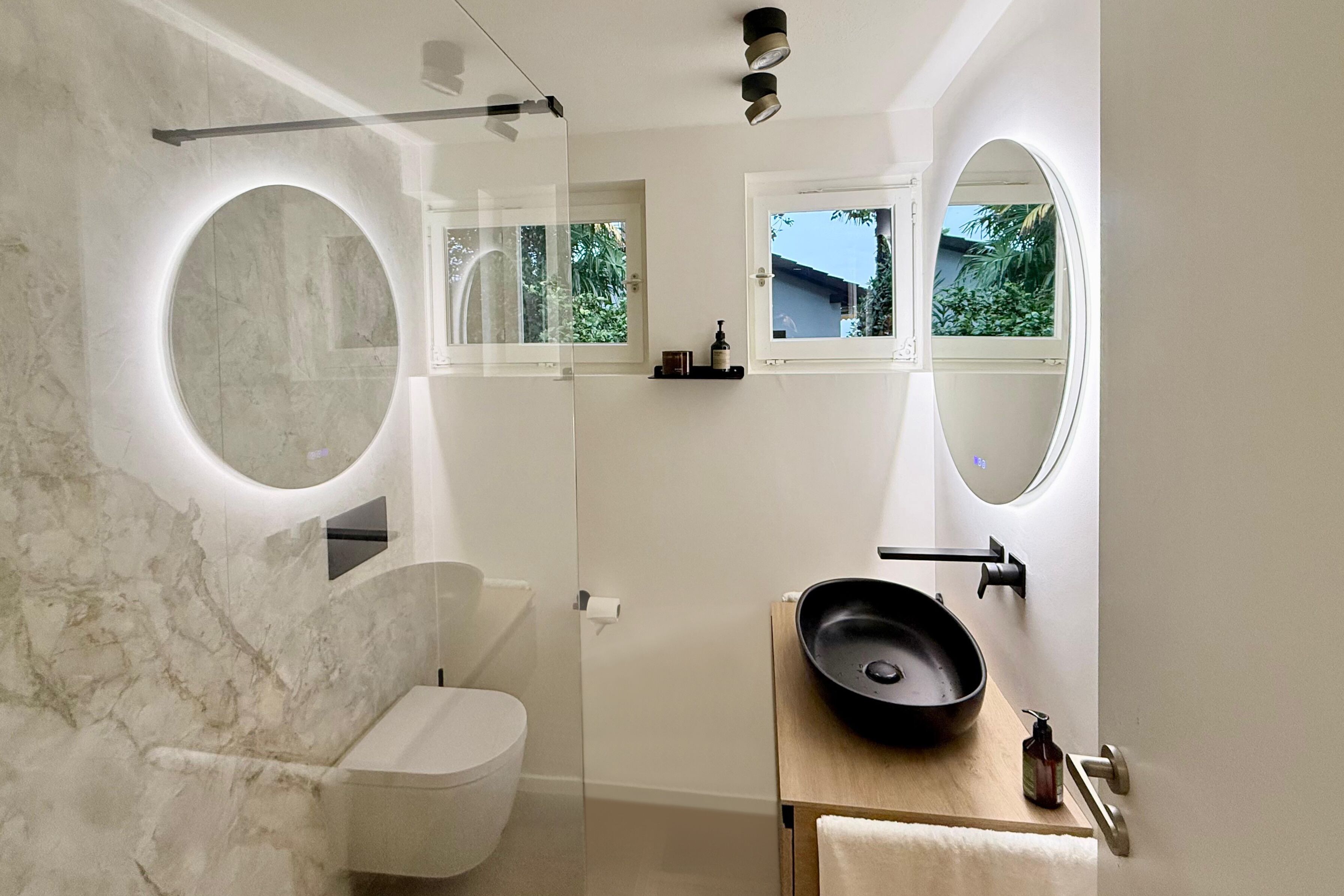 Een moderne badkamer met toilet, een wastafel met een zwart wasbakje, en een spiegel met ronde lijst.