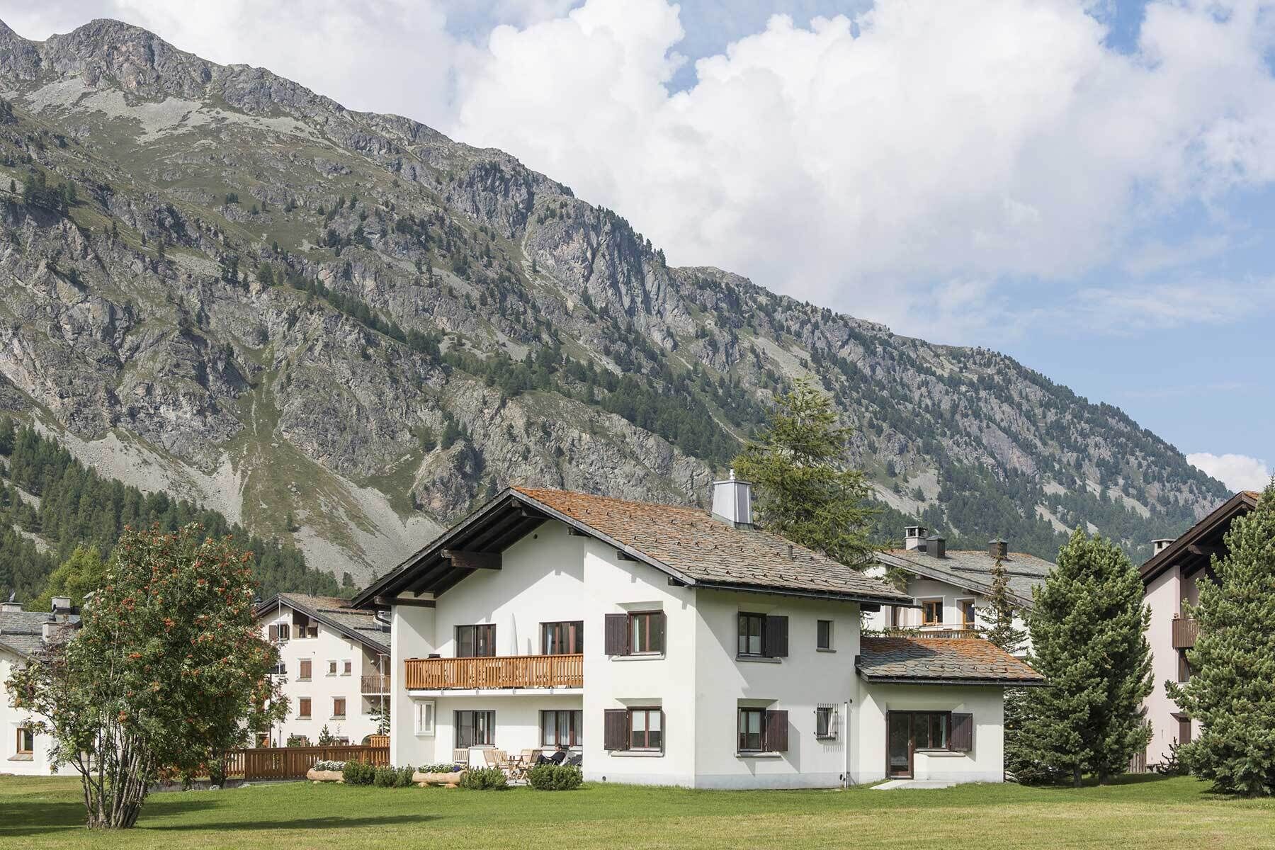 A two-story building with a balcony, surrounded by a grassy area and a mountain backdrop.