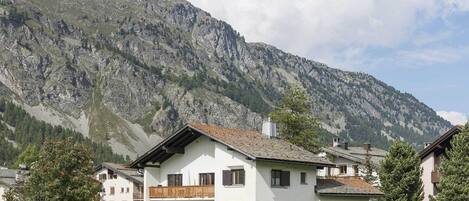 A two-story building with a balcony, surrounded by a grassy area and a mountain backdrop.