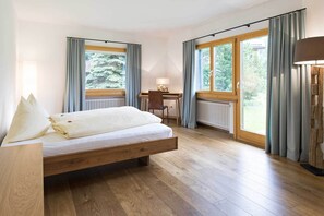 Ein Schlafzimmer mit einem hölzernen Bett, einem Schreibtisch und einem Stuhl, großen Fenstern mit Vorhängen und Blick ins Grüne.