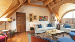 Una habitación estilo loft acogedora, con techo alto, vigas de madera, un sofá blanco, sillones azules, un tapete estampado y una mesa de centro de madera.