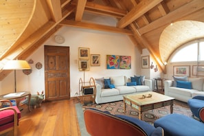 Ein gemütlicher Loftsraum mit hoher Decke, Holzbalken, einem weißen Sofa, blauen Sesseln, einem gemusterten Teppich und einem Holztisch.
