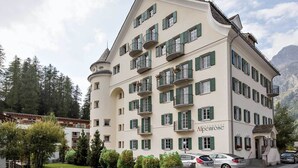 Un hotel de varios pisos con balcones, un cartel que dice 'Alpenrose' y un paisaje montañoso de fondo.