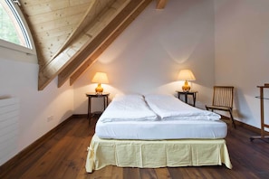 Ein Zimmer mit schrägem Dach, zwei Betten mit weißen Bettwäsche, zwei Nachttischlampen, ein Holzstuhl und ein Fenster.