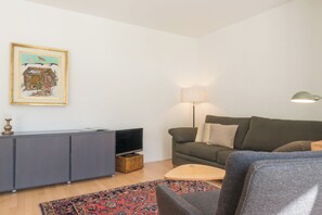 Ein Wohnzimmer mit grauem Sofa, einem kleinen runden Tisch, einem Fernseher auf einer Kommode, einer Lampe und einem farbenfrohen Teppich.