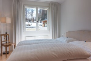Ein Schlafzimmer mit Bett, Stuhl, einem Fenster mit Vorhängen und Blick auf eine verschneite Landschaft.