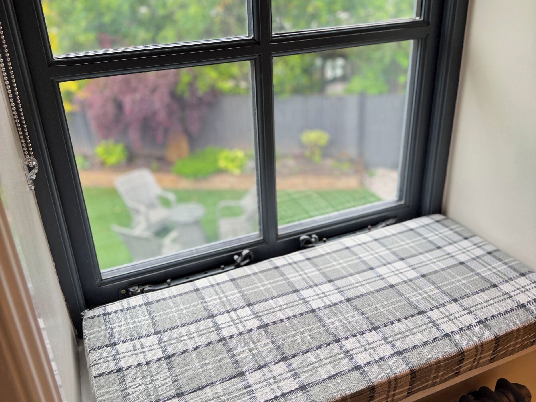 Un siège de fenêtre avec un coussin à carreaux, une vue sur un jardin et une chaise blanche à l’extérieur.