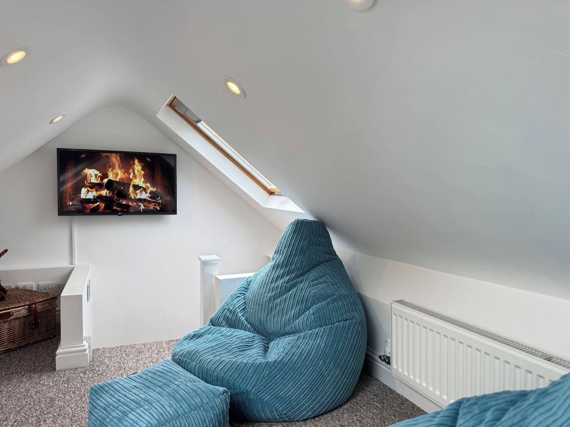 Acogedor dormitorio en ático con techo inclinado, un televisor que muestra una chimenea, un sillón de peluche azul y un radiador blanco.