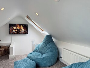 Ein gemütliches Schlafzimmer im Dachgeschoss mit schrägen Wänden, einem Fernseher, der einen Kamin zeigt, einem blauen Sitzsack und einer weißen Heizung.