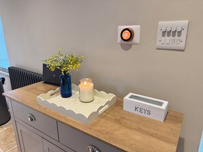 Una habitación con mostrador de madera, un florero con flores, una vela encendida, un termostato y una caja para llaves.