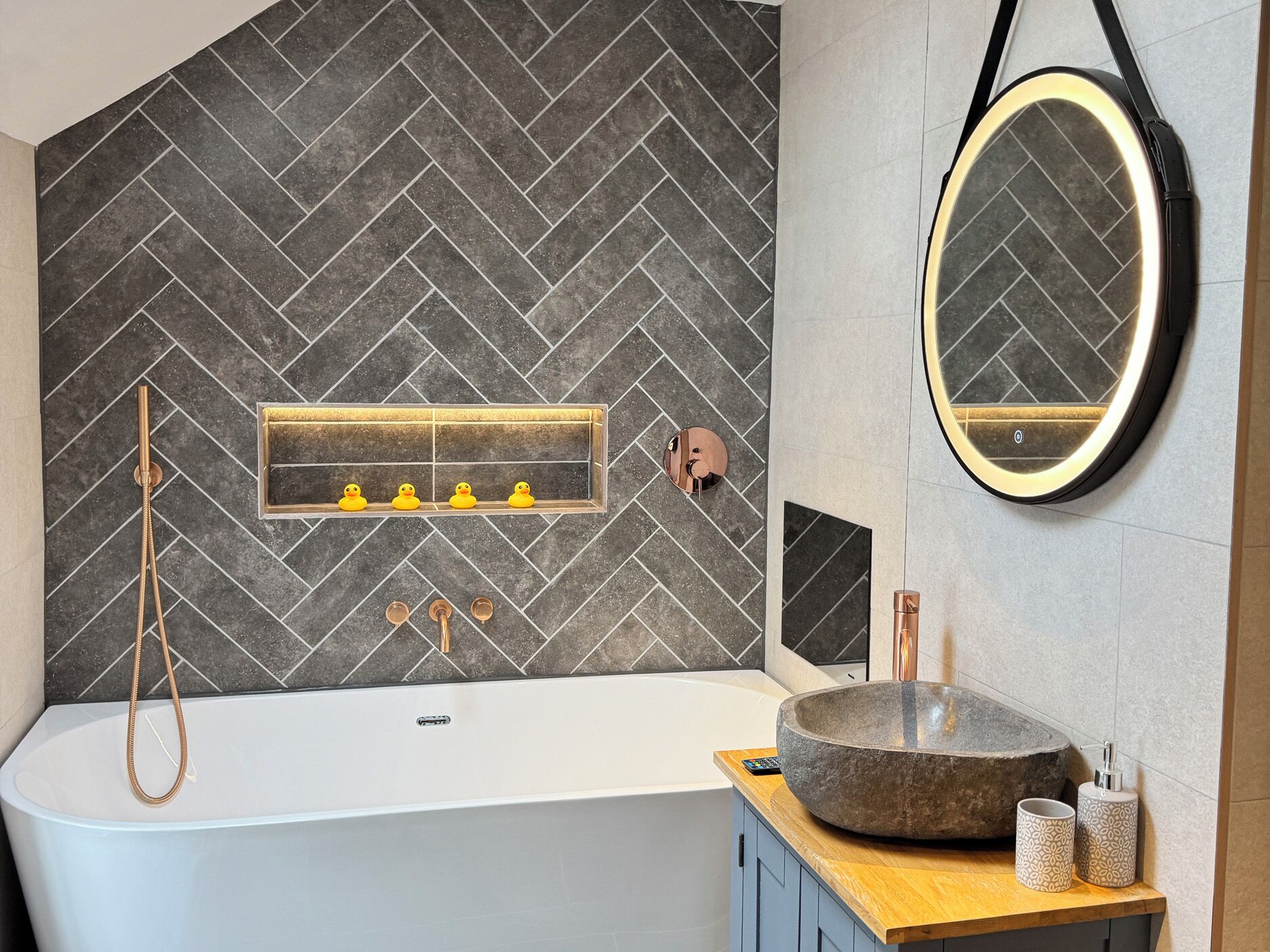 A modern bathroom with a herringbone tile backsplash, a freestanding bathtub, a round mirror, and a wooden vanity with a stone basin.