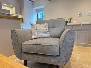 A grey upholstered armchair with a plaid pillow in a modern kitchen with grey cabinetry and a tiled floor. - Railway Cottage (Oswestry)