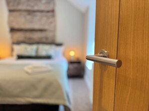 A wooden door with a silver handle, a stone wall, and a bed with a white bedspread in the background. - Railway Cottage (Oswestry)