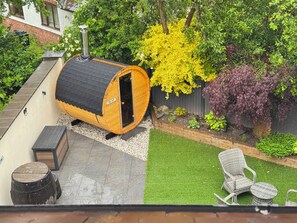 A sauna with a chimney, surrounded by a garden with various plants and a patio area with outdoor furniture. - Railway Cottage (Oswestry)