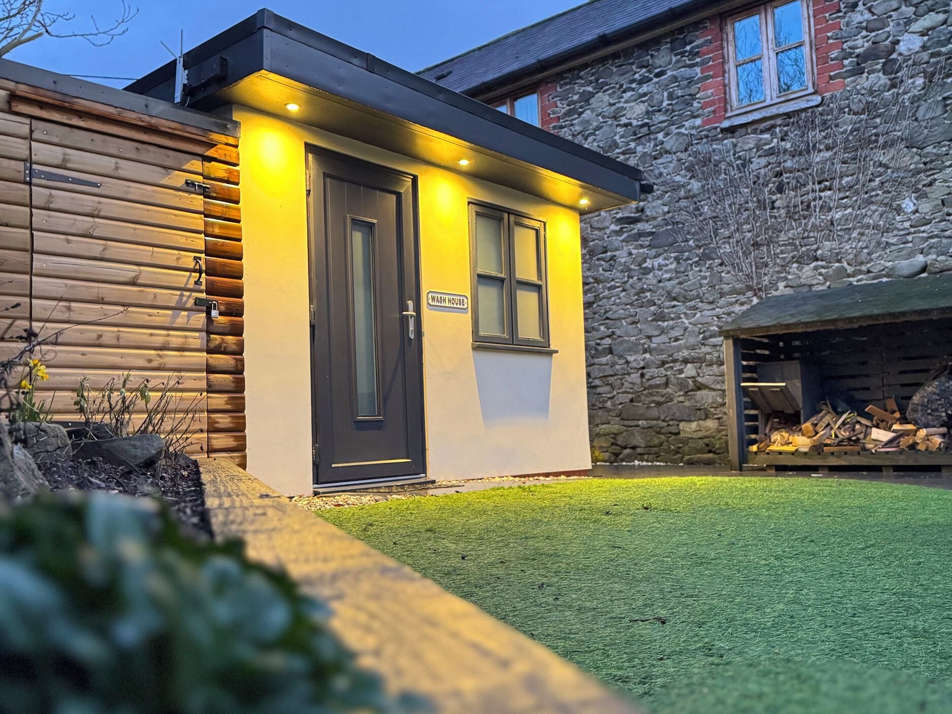A small, modern building with a dark door and windows, labeled 'WASH HOUSE,' adjacent to a stone wall and a grassy area.