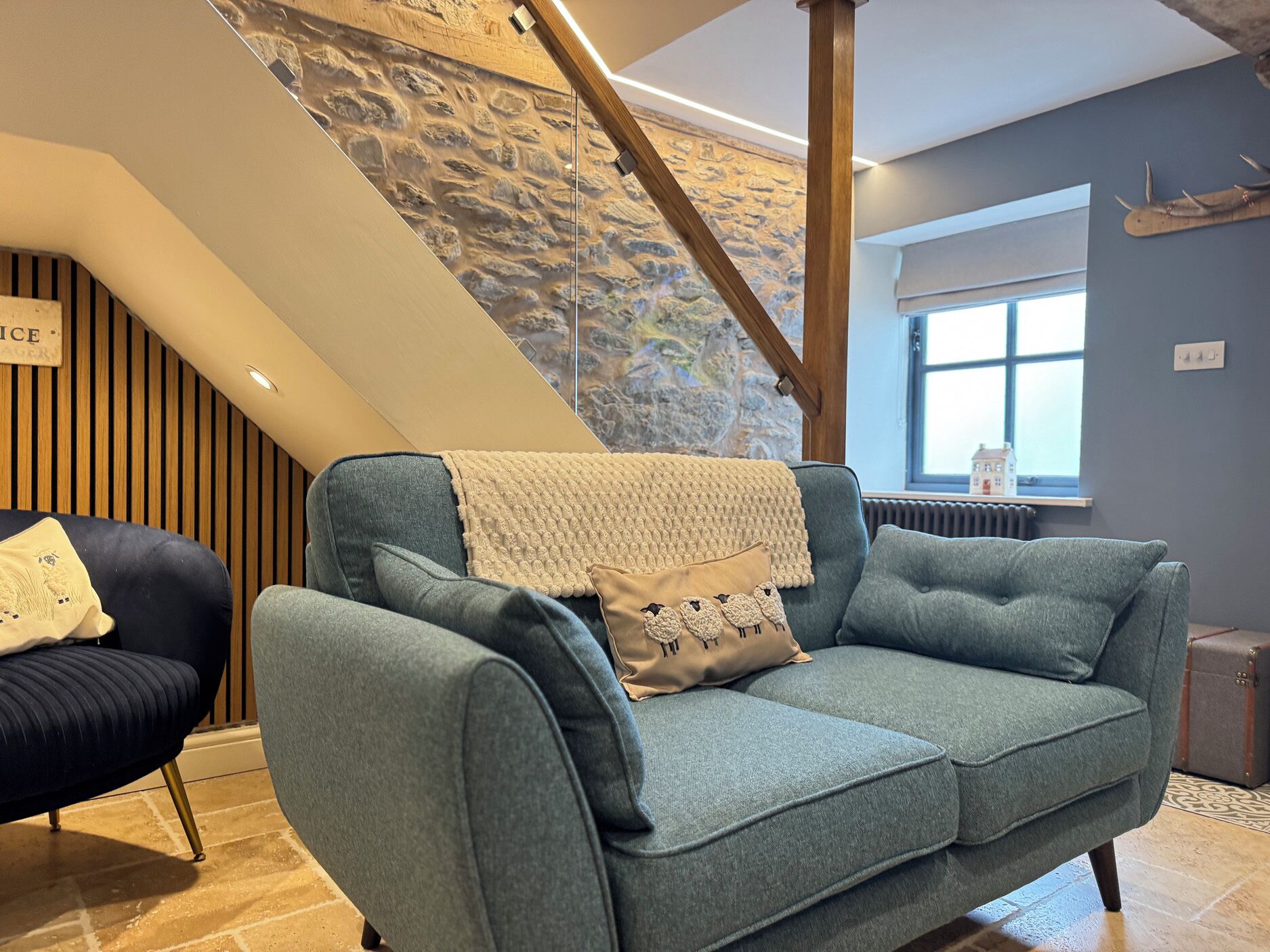 A modern living room with a blue sofa, a wooden chair, a stone wall, and a staircase.